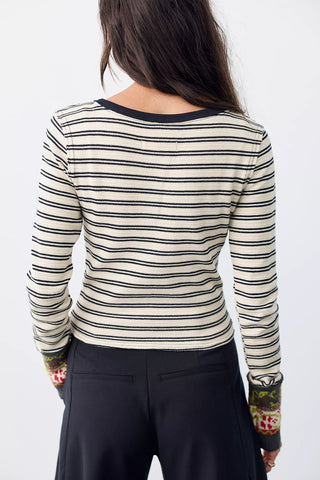A woman shown from the back wearing a cream ribbed long-sleeve top with thin black horizontal stripes and patterned cuffs, paired with black pants, against a plain white background.