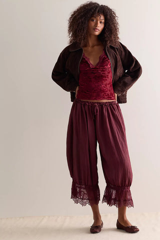A model wearing a dark brown jacket over a deep burgundy velvet camisole and matching pleated harem-style pants with lace trim, paired with dark brown flats, against a plain off-white studio background.