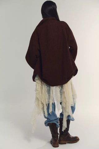A person shown from the back wearing an oversized dark brown knit cardigan over a layered outfit with distressed cream lace, blue jeans, and brown leather boots, photographed against a plain light gray studio background.