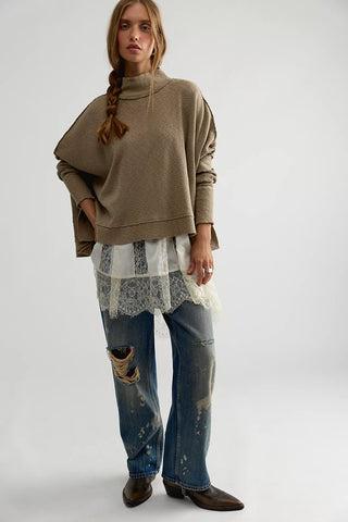A model stands against a light gray studio background wearing a taupe-brown cropped turtleneck sweater, a white lace-trimmed shirt, distressed blue jeans, and dark brown ankle boots.