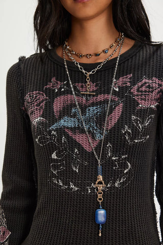 A close-up of a person wearing a black textured knit top with a pink and blue rose graphic, layered with multiple silver and gold necklaces, including a long pendant necklace with a blue bead.