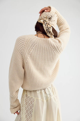 A woman shown from the back wearing a cream-colored chunky knit sweater, a cream lace skirt, and a patterned beige and black headscarf, set against a plain light gray studio background.