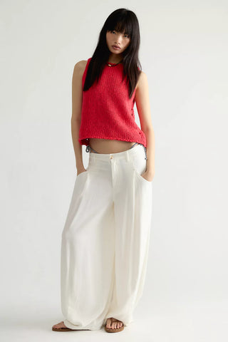 Woman wearing a red sleeveless top and white pants on a plain background