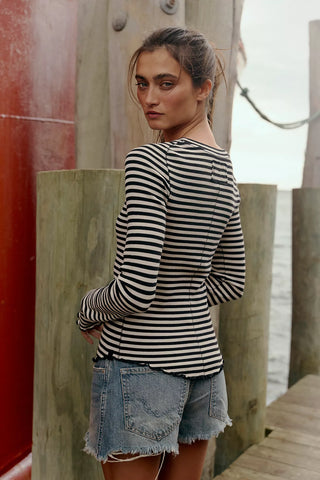 Woman wearing a black and white striped shirt and denim shorts standing against a wooden background.