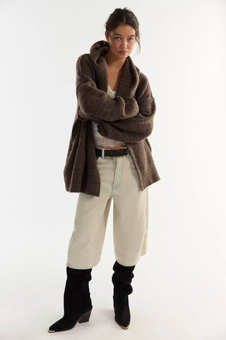 Women wearing a brown knit cardigan and cream jeans in front of a neutral photo background.