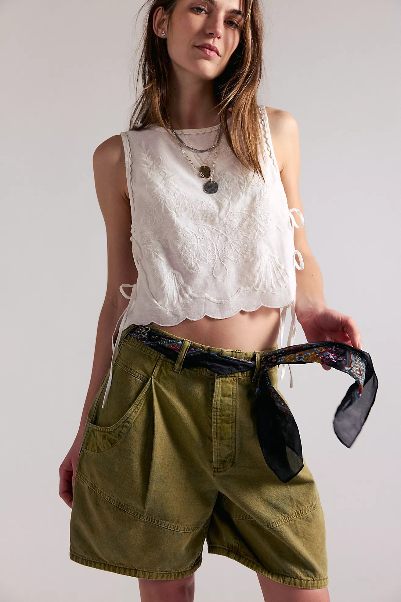 Woman wearing a white sleeveless top and green shorts, holding a black belt against a plain background.