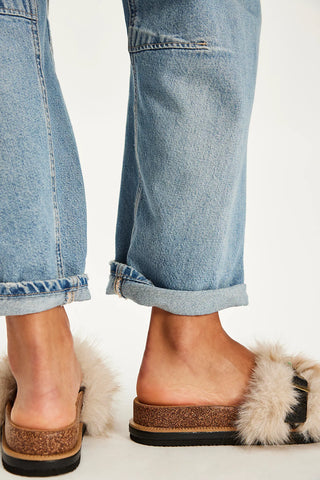 Close-up of a person wearing light blue cuffed jeans and brown slide sandals with fluffy cream trim, shown against a plain light background.