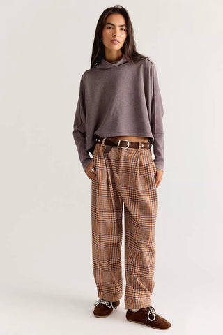 A model stands against a light gray studio background wearing a cropped taupe-brown cowl-neck sweater, high-waisted brown and orange plaid trousers with a brown belt, and brown suede loafers.