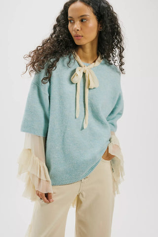 Woman wearing a light blue sweater with a beige bow tie and beige pants on a white background