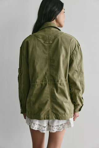 A woman shown from the back wearing an olive green utility jacket over a white lace-trimmed skirt, photographed against a plain light gray studio background.