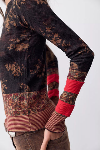 A close-up of a person wearing a long-sleeve knit sweater with a dark brown and rust floral pattern, contrasting red ribbed cuffs, and a patterned hem, paired with brown pants against a light gray background.