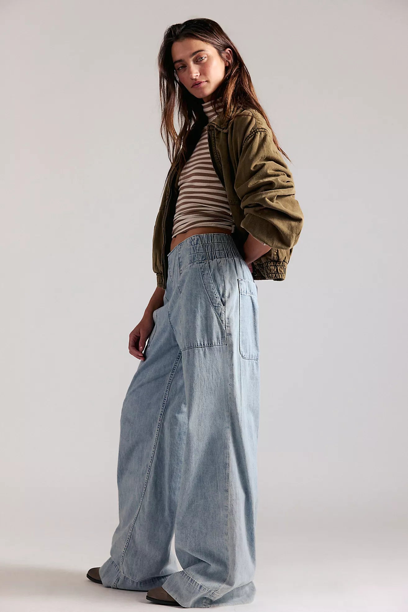 Woman wearing a green jacket, striped shirt, and light blue jeans on a plain background