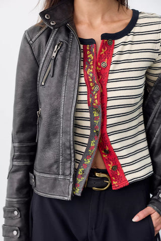 A close-up of a person wearing a dark gray distressed leather moto jacket over a cream and black striped knit top with a red floral button-front panel, paired with dark pants and a black belt.