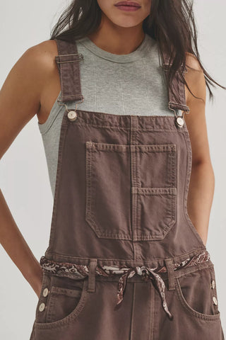 Person wearing brown overalls over a light gray tank top against a neutral background