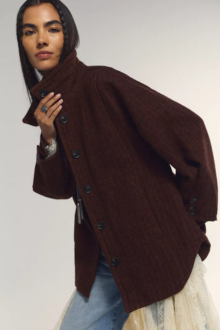 A woman wearing a dark brown wool-blend coat with a high collar and dark buttons, paired with light blue jeans and a cream lace skirt, photographed against a neutral light gray background.