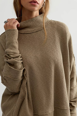 A close-up of a woman wearing a ribbed knit turtleneck sweater in a warm taupe-brown color, with her hand adjusting the collar.