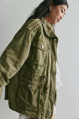 A woman wearing an oversized olive green military-style utility jacket with multiple flap pockets and silver snap buttons, photographed against a plain light gray studio background.