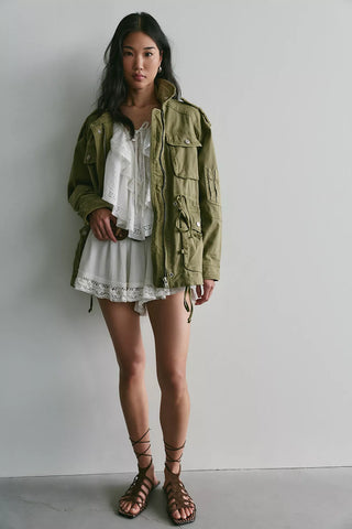 A woman stands against a plain light gray wall wearing an olive green military-style jacket over a white ruffled top and white skirt, with brown lace-up sandals.