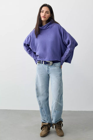 A woman modeling a loose, oversized lavender-purple turtleneck sweater, paired with light-wash blue jeans, a silver belt, and brown suede boots, standing against a plain light gray studio background.