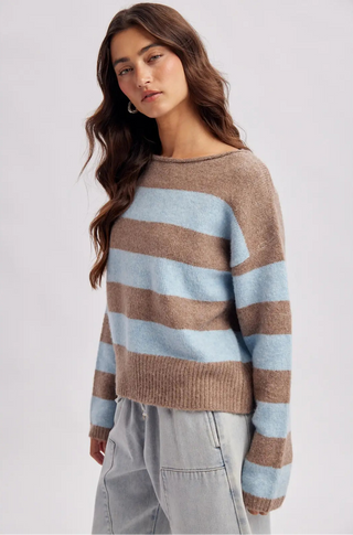 A woman wearing a fuzzy knit sweater with horizontal stripes in light blue and brown, paired with light gray pants, photographed against a plain light gray studio background.