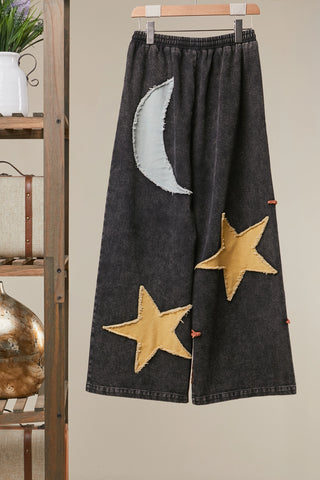 Dark denim pants with star and moon patches on a hanger against a neutral wall.