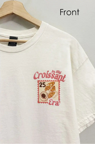 An off-white short-sleeve t-shirt hanging on a wooden hanger against a light gray wall. The shirt features a graphic print on the left chest with the text 'In My Croissant Era' and a vintage-style croissant illustration inside a red stamp-like border with the number '25'.