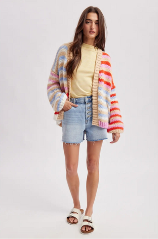 Woman wearing a colorful striped cardigan, yellow top, denim shorts, and sandals on a white background