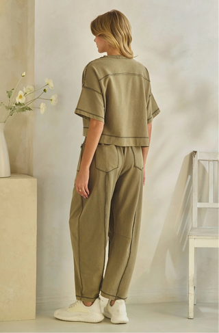 A woman stands with her back to the camera wearing a matching two-piece outfit consisting of a short-sleeve cropped top and wide-leg pants in a muted olive khaki color. She is wearing white sneakers in a minimalist room with a white chair and a vase of flowers.