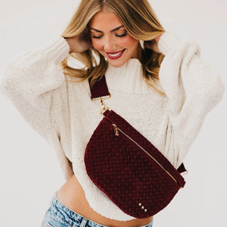 A woman wearing a cream knit sweater and blue jeans models a burgundy woven crossbody bag with gold hardware against a light gray background.