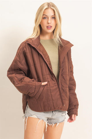 A blonde woman wearing a brown quilted zip-up jacket over a beige ribbed top and light denim shorts, standing against a plain light beige studio background.