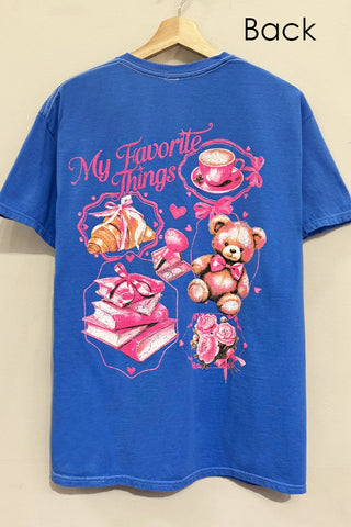 A blue short-sleeve t-shirt hanging on a wooden hanger against a light beige wall. The back of the shirt features a large graphic print with pink text reading 'My Favorite Things' and illustrations of a teddy bear, books, a croissant, a cup of coffee, and roses.