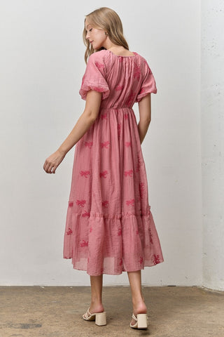 Woman wearing a pink dress with butterfly patterns against a white wall.