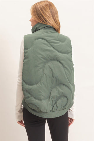 A woman shown from the back wearing a sage green quilted puffer vest over a white long-sleeve top and black pants, against a plain white studio background.