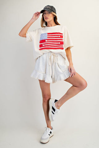 Woman wearing a white t-shirt with an American flag design, white shorts, and a cap on a plain background