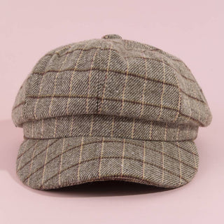 A structured flat cap in a textured herringbone pattern of gray and beige, featuring a subtle windowpane grid design, set against a solid light pink background.