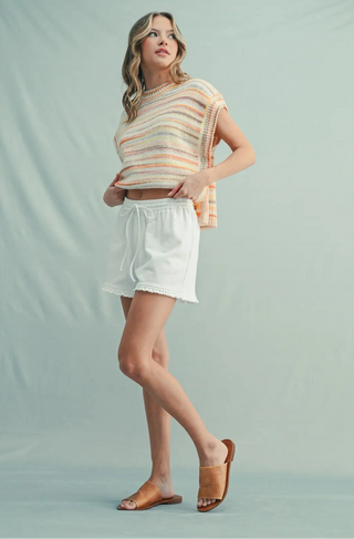 A woman modeling a short-sleeve knit top with horizontal pastel stripes in beige, orange, and white, paired with white shorts and tan slide sandals, against a light blue-green studio backdrop.