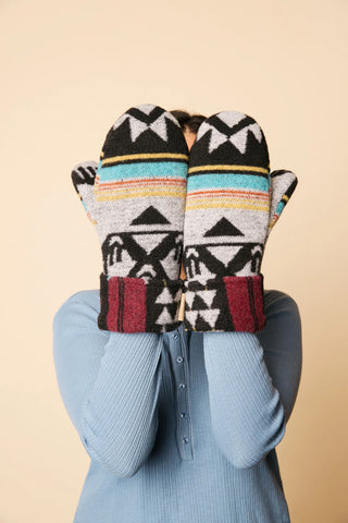 A person wearing a light blue ribbed long-sleeve top holds a pair of patterned knit mittens in front of their face. The mittens feature a Southwestern geometric design in black, white, gray, turquoise, yellow, and burgundy on a solid beige background.