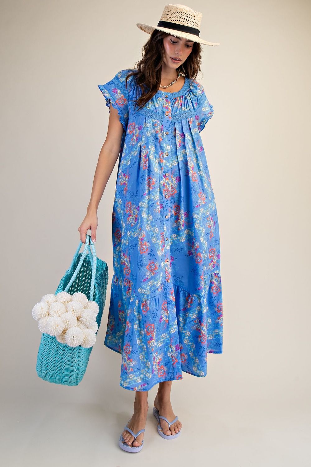 Woman wearing a blue floral dress holding a blue bag with white pom-poms against a plain background