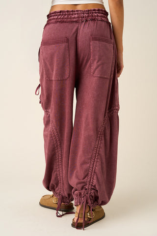 A person shown from the back wearing loose, wide-leg drawstring pants in a dusty mauve or burgundy tone, paired with tan sandals, against a plain light beige studio background.