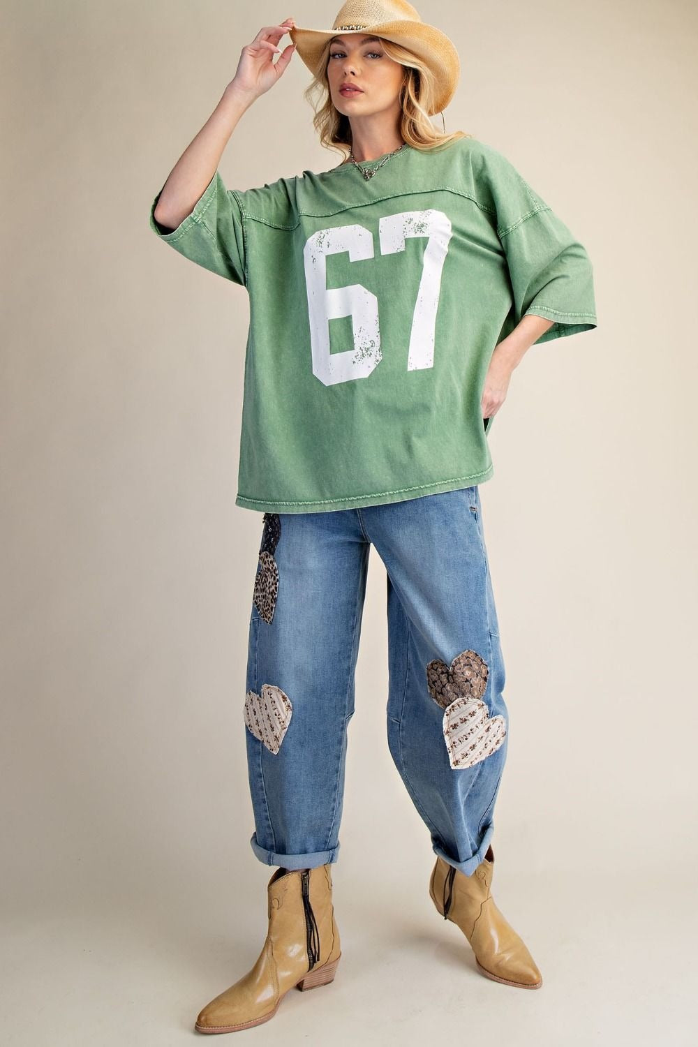 Woman wearing a green oversized shirt with the number 67, blue jeans with patches, and tan boots on a beige background.