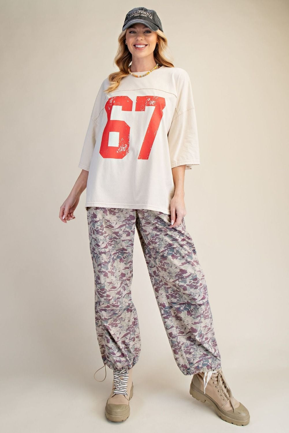 Woman wearing a white oversized shirt with red number '67', floral pants, and beige shoes on a plain background