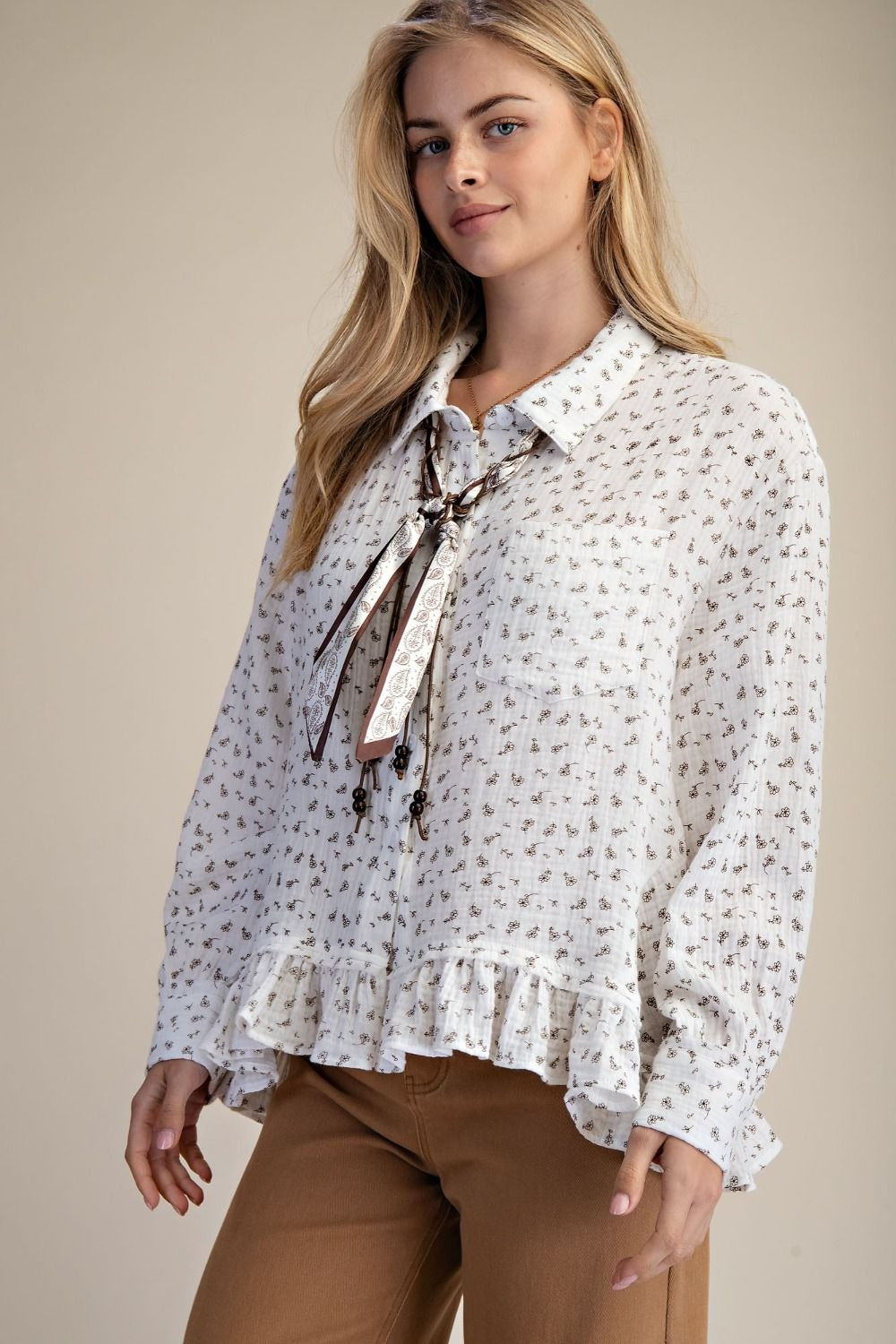 Woman wearing a white floral blouse with a tie and brown pants on a beige background