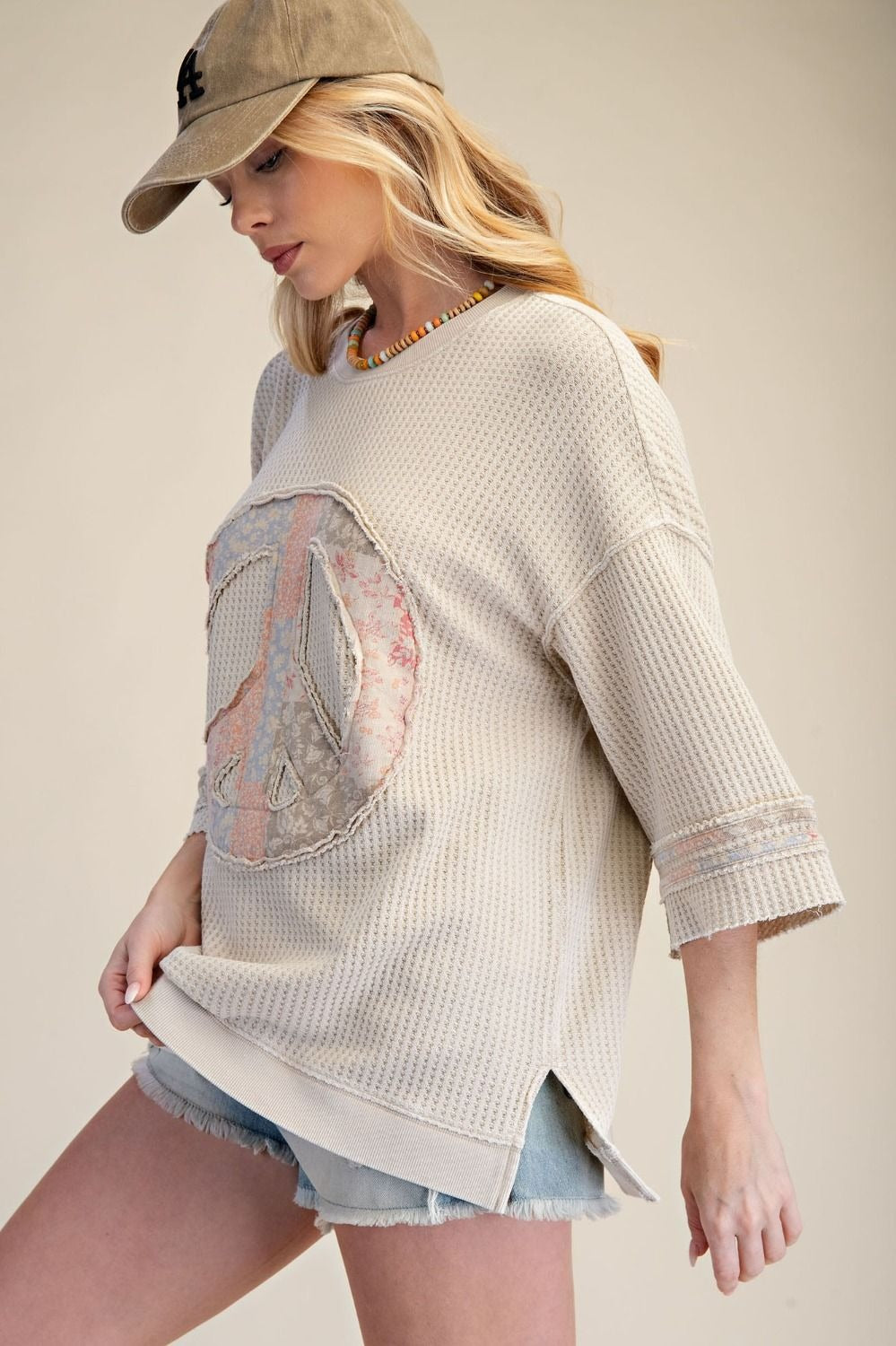 Woman wearing a beige sweater with a peace sign design, denim shorts, and a cap on a neutral background