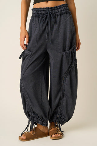A person wearing dark charcoal gray baggy drawstring pants with side cargo pockets and elastic ankle cuffs, paired with brown sandals against a plain light beige studio background.