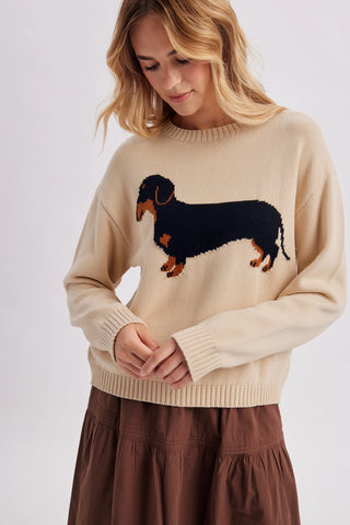 A woman wearing a beige crewneck sweater with a large black and brown dachshund graphic on the chest, paired with a brown skirt, against a light gray studio background.