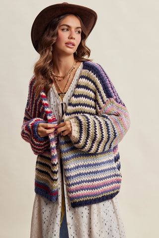 A woman wearing a chunky, multicolored striped knit cardigan, a brown wide-brim hat, and a cream-colored dress, posed against a solid light beige background.