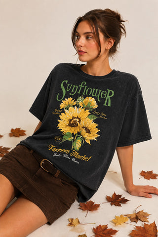 A woman wearing a faded black graphic t-shirt with a large yellow sunflower print and green text, paired with brown corduroy shorts, posed on a white floor scattered with autumn leaves.