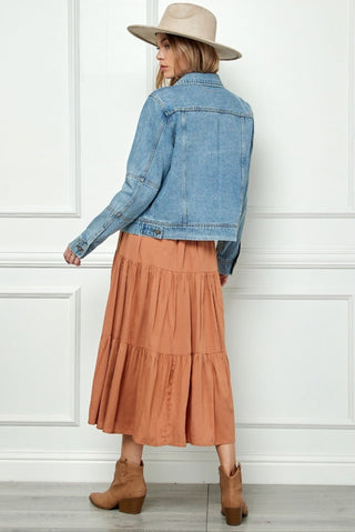 Woman wearing a denim jacket, tan hat, and tiered brown skirt against a white paneled wall.