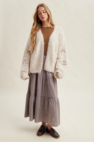 Woman wearing a textured cardigan, brown top, and plaid skirt on a white background