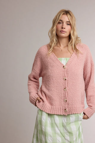 A woman wearing a fuzzy light pink button-up cardigan over a light green and white gingham dress, standing against a plain light gray studio background.